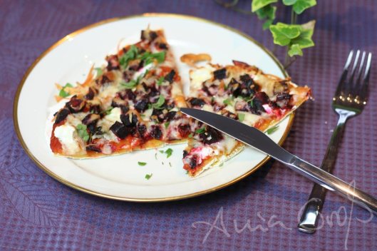 Lila Untergrund, mittelgroßer Tller mit Goldrand, darauf zwei Ecken Pizza. Man sieht Besteck und ein paar Efeublätter. Foto: © Ania Groß